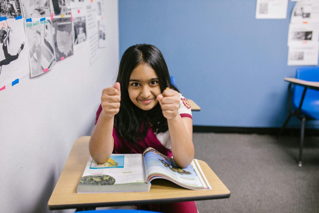 A confident young girl in a classroom giving a thumbs-up while reading a book.